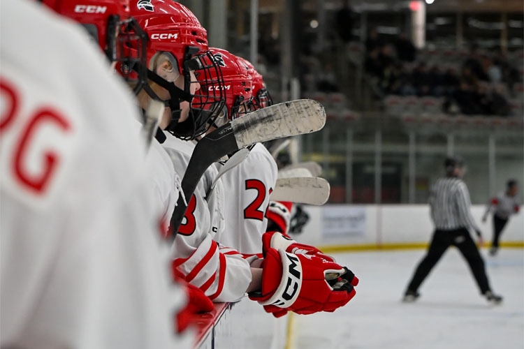 Trojans women's hockey players in red helmets and white jerseys lean over the bench while holding their sticks, with a referee skating on the ice in the background.