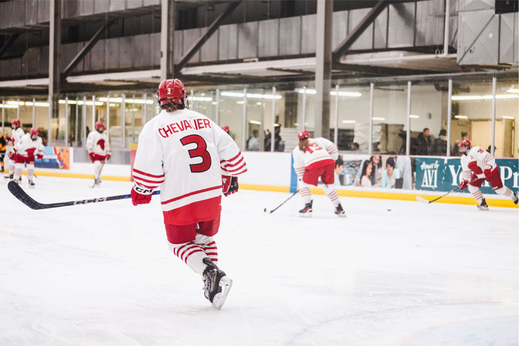Trojans women's hockey players in red-and-white uniforms skate during warmups on an indoor ice rink, with one player wearing jersey number 3 in the foreground.