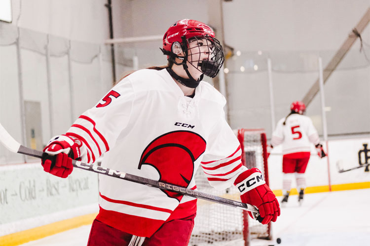 Trojans women's hockey player in a white and red uniform and red helmet holds a stick on an indoor ice rink, with another player and the goal in the background.