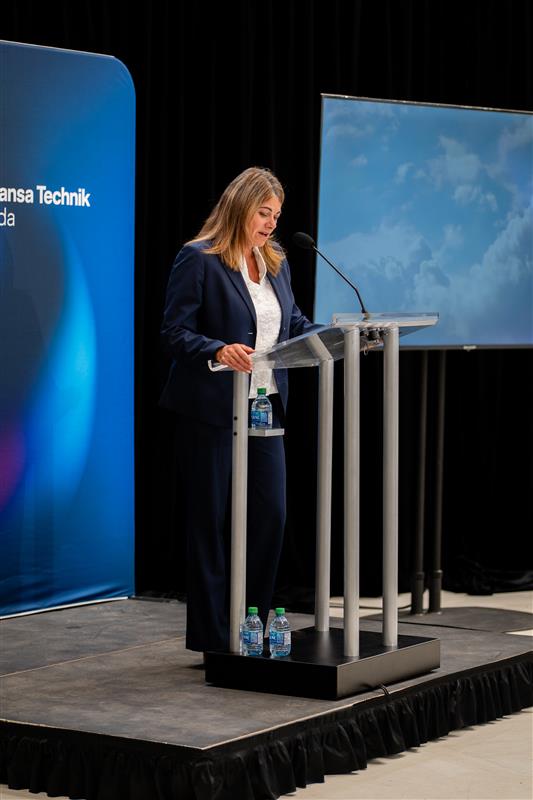 A woman in a navy suit delivers a speech at a podium during a corporate event, with a branded backdrop and a screen displaying a cloudy sky.