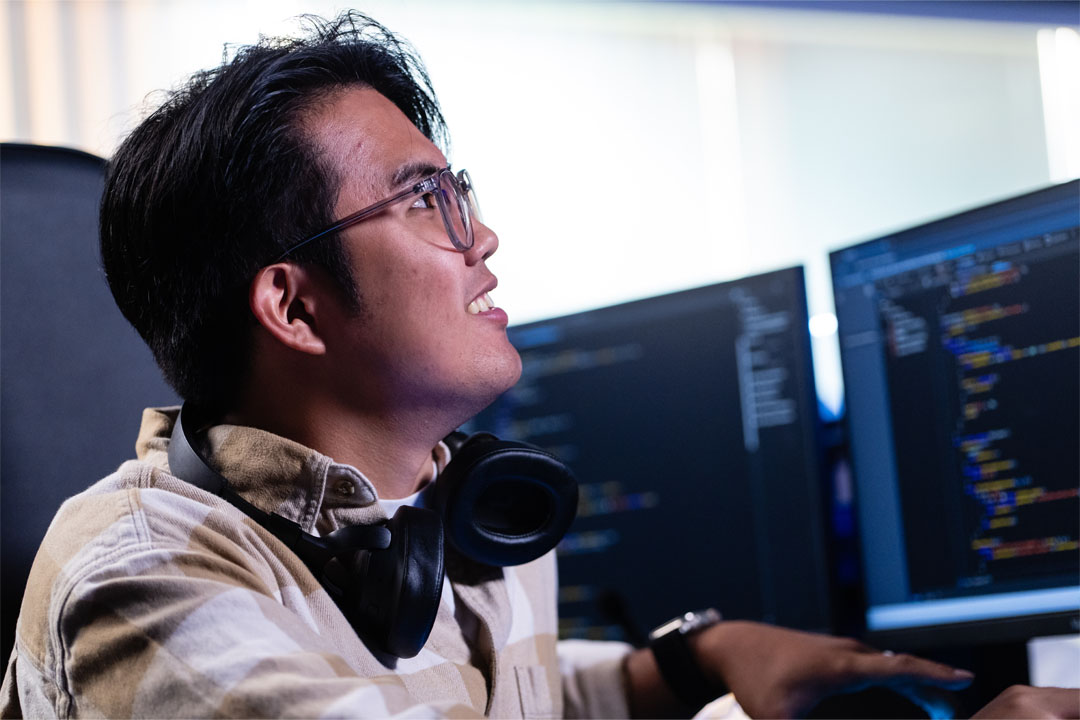 A young man wearing glasses and headphones, smiling while engaging with dual computer monitors displaying code.
