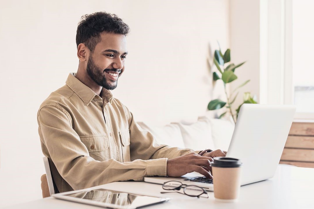 A man in a tan, collared shirt sits at his desk and smiles at his laptop.