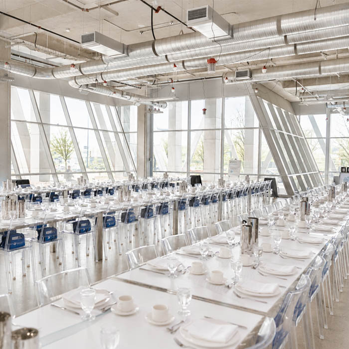 Bright glass‑walled event hall with long banquet tables and clear chairs.