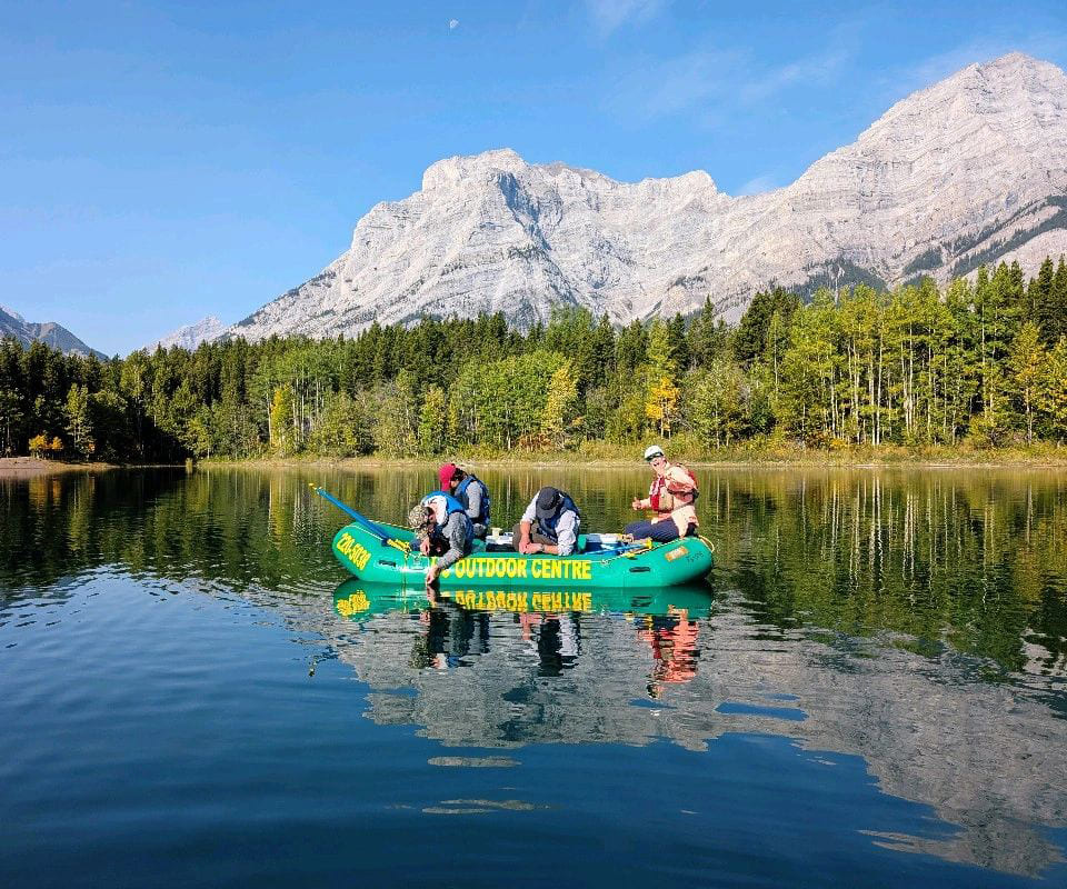 Group of people in an inflatable boat on calm water, surrounded by mountains and trees engaged in outdoor activities.