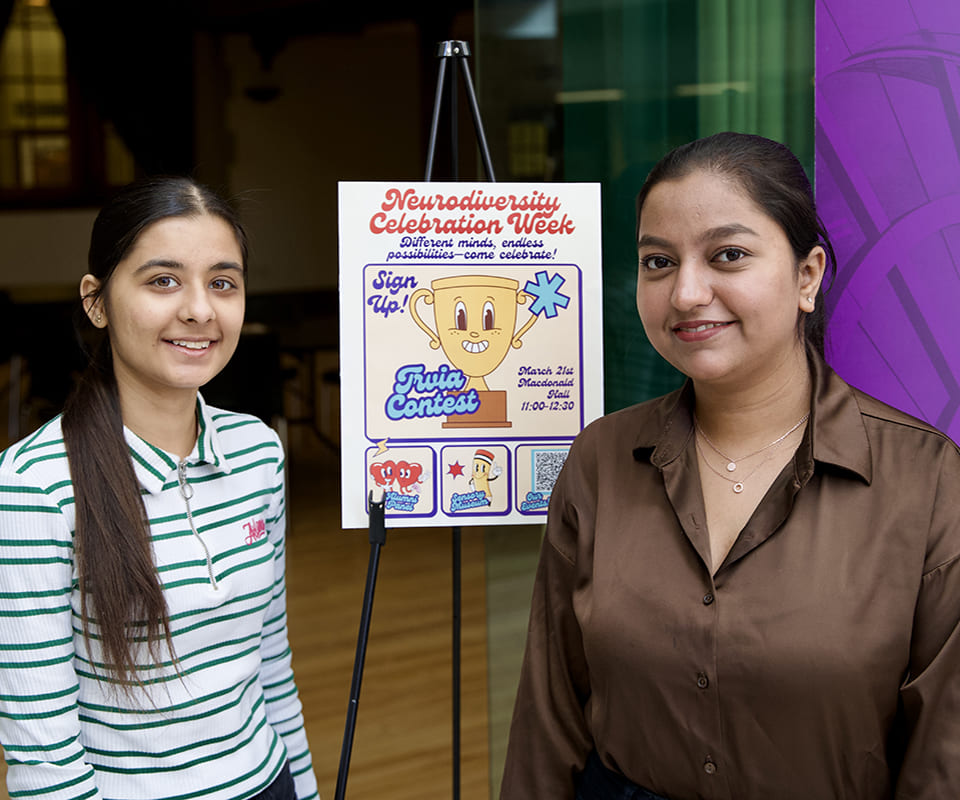 Two young women standing next to a sign promoting the Neurodiversity Celebration Week trivia contest.