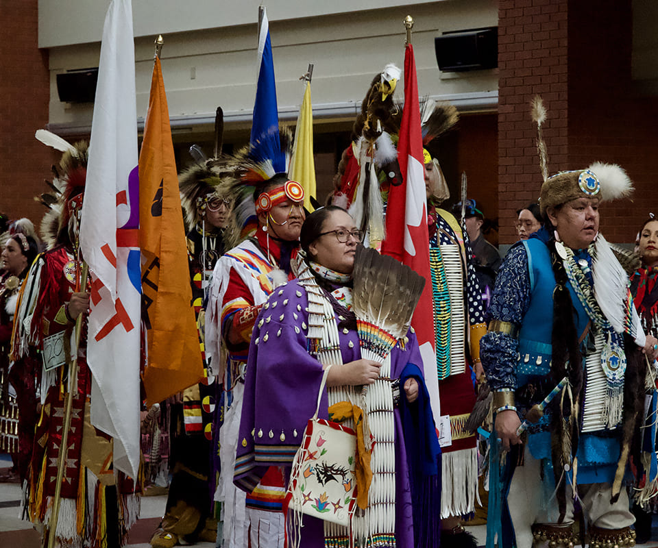 Indigenous participants in traditional attire at a cultural event, holding flags and ceremonial items.