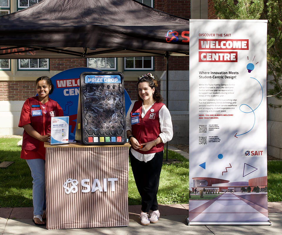 Two smiling women stand by a Welcome Centre booth at SAIT featuring a prize drop game and an informative banner about student services.
