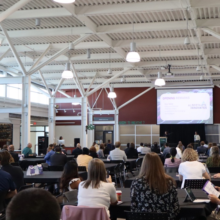 Audience seated at tables in a large event space – facing a stage with a presentation screen displaying opening remarks for an Alberta Innovates symposium.