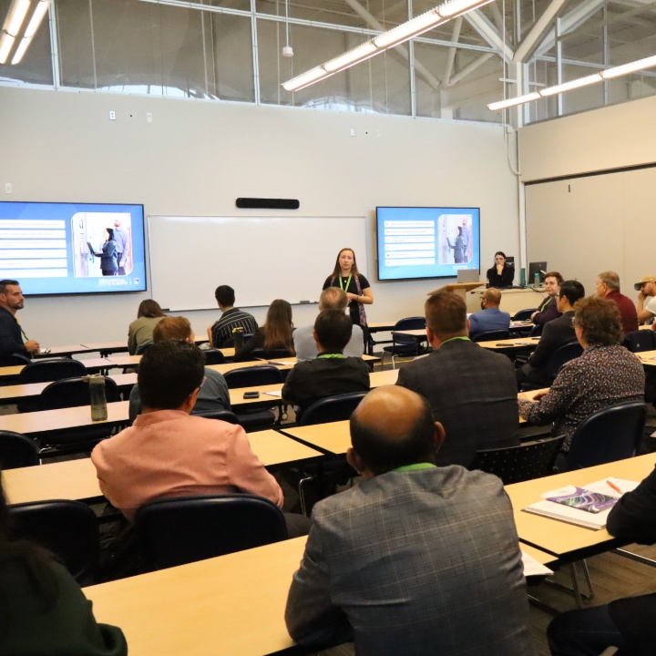 Presenter speaking at the front of a classroom – audience seated at tables facing slides displayed on two screens.