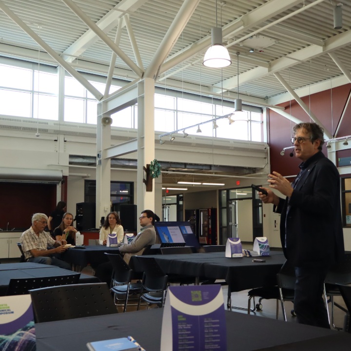 Presenter speaking in a large event space – small groups seated at round tables listening, with signage and materials visible on tables.