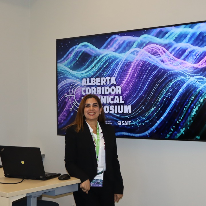Person standing beside a desk with a laptop in front of a screen displaying “Alberta Corridor Technical Symposium” with a colourful wave graphic and SAIT branding.