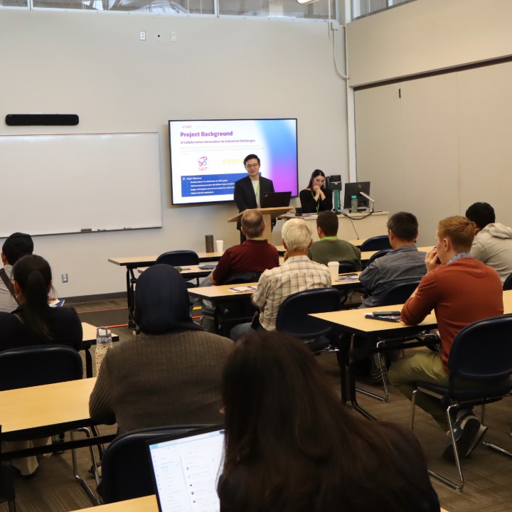 Two presenters at the front of a classroom – audience seated at tables facing a slide titled “Project Background,” with SAIT branding displayed on the screen.
