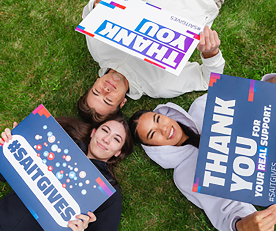 Three young adults lying on the grass, holding signs that say Thank You and SAITGIVES to express gratitude and support.