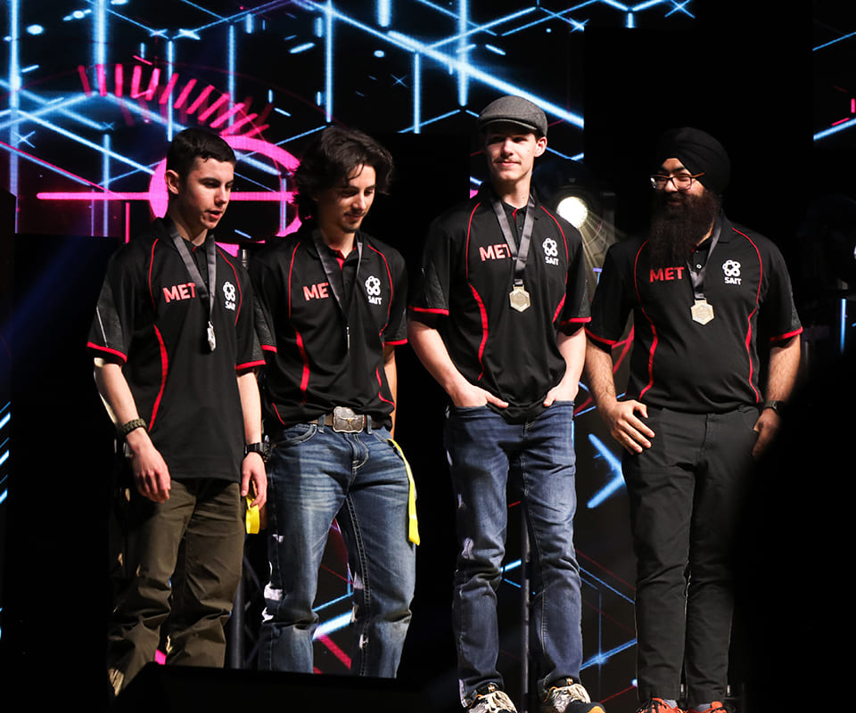 Four individuals wearing matching black polo shirts with MET printed on them standing on stage during a robotics competition event.