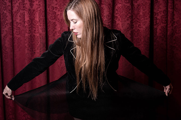 female Caucasian with long brown hair wearing black outfit in front of a red curtain like those at a theatre