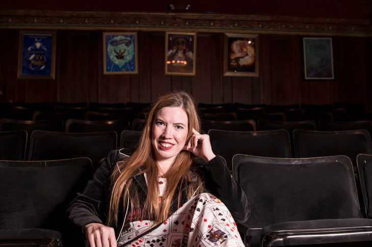female Caucasian sitting in audience seat of theatre, long brown hair, wearing a black top and skirt with playing card images on it.