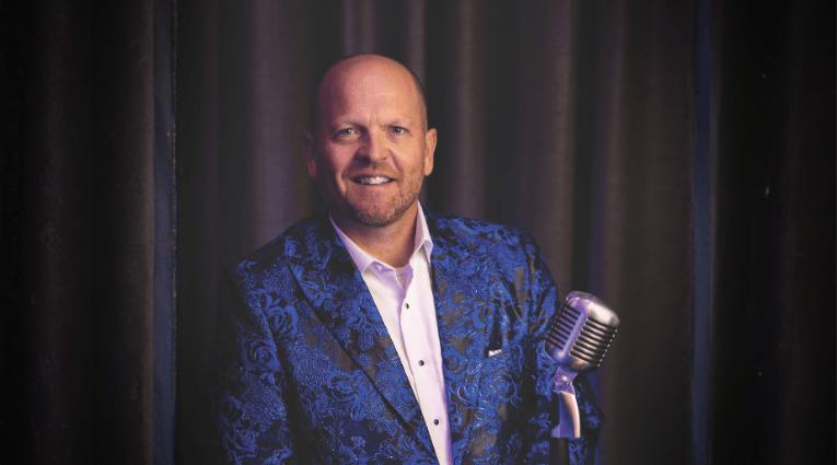 white male wearing a blue patterned blazer standing at old mic