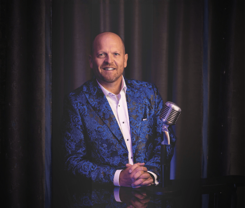 white male wearing a blue patterned blazer standing at old mic