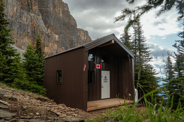 Small brown cabin with Canadian flags by the door, surrounded by evergreen trees and rocky mountains under a cloudy sky.