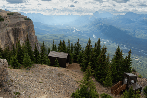 Mountain overlook with small cabins and evergreen trees, looking out over a broad valley with a winding river under a cloudy sky.