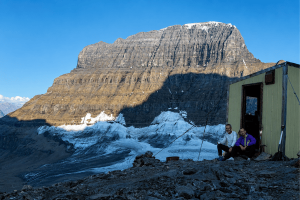 Two hikers rest beside a small mountain hut, overlooking a glacier and steep rocky peaks under a clear blue sky.