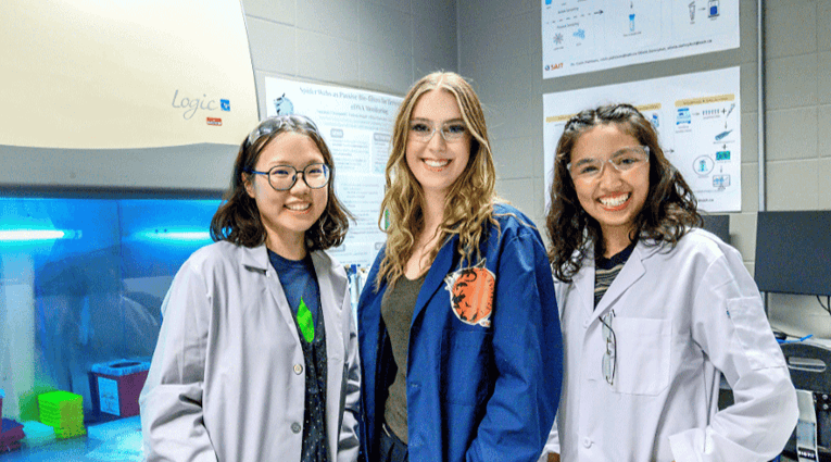 Three people in lab coats posing in a lab with equipment and posters in the background.