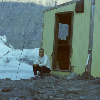 Person beside a small cabin overlooking a glacier.