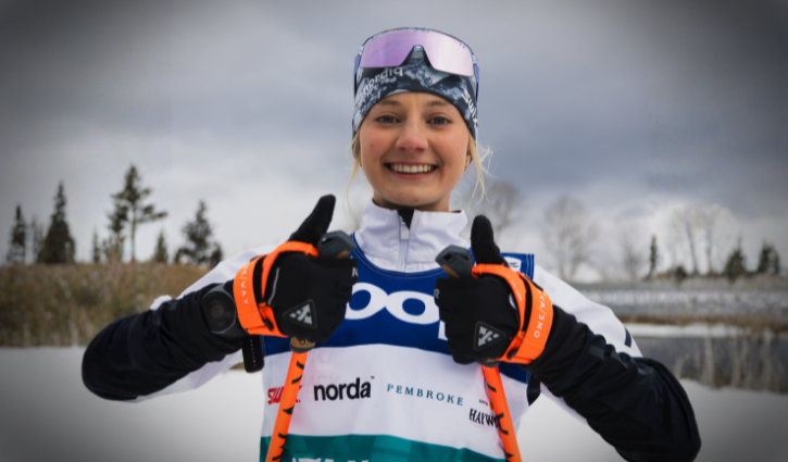 Young white woman in competitive ski gear with two thumbs up.