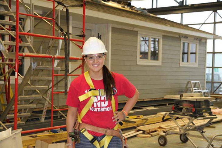 A student wearing a hard hat, safety gloves and vest poses in front of a home build in progress inside the Home Builders lab on SAIT campus.