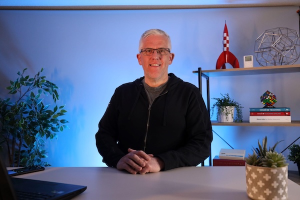 Man smiling at a desk in a home office with shelves, books, and plants in the background.