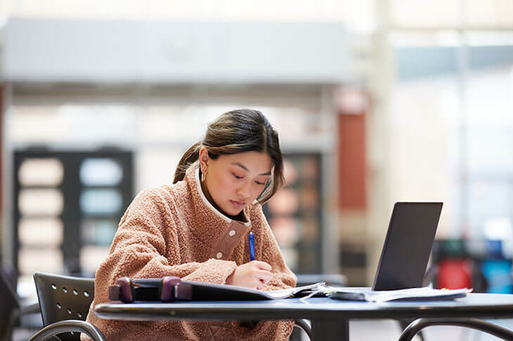 A young woman sits at a table working with a notebook and laptop, in a bright room.