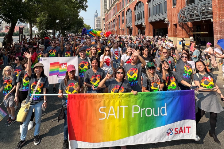 People wearing t-shirts with the rainbow SAIT logo standing behind a banner that says 'SAIT Proud'
