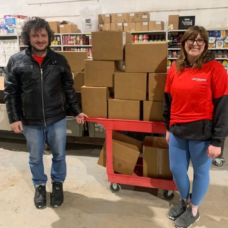 Two people smile while standing in front of cardboard boxes stacked on a cart.