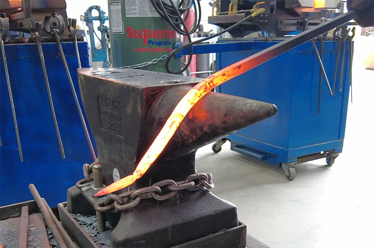 A glowing metal rod sits on an anvil in a blacksmith shop. In the background, there’s a propane tank and a blue cart with tools. 