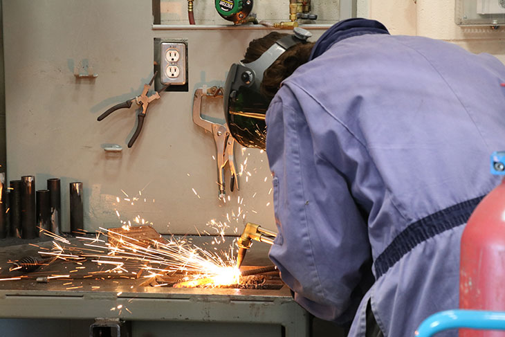 A student in a welding mask cutting metal. There are bright orange and yellow sparks flying.