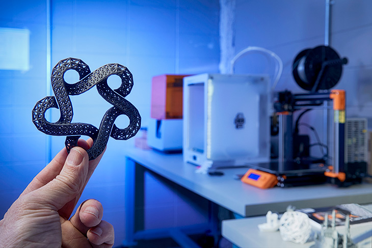 hand holding a 3d printed SAIT catalyst logo with 3d printers blurred in the background