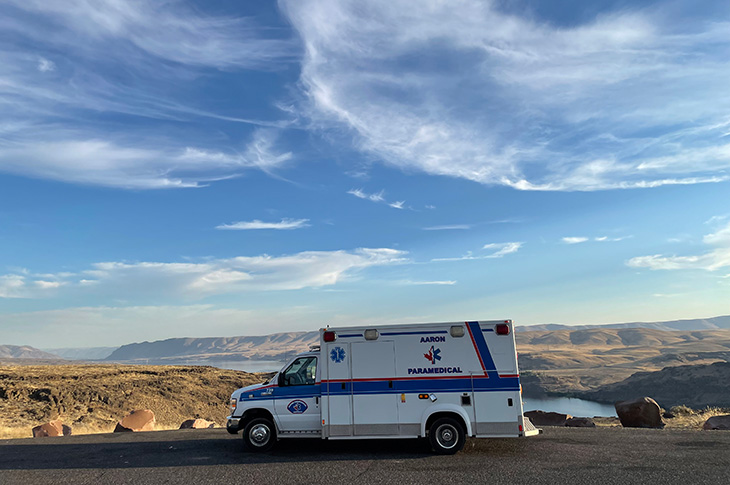 An ambluance is parked on the side of the road with a river and blue sky in the background. The ambulance has an 'Aaron Paramedical' logo on it.