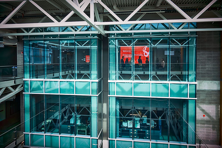 Inside SAIT’s Aldred Centre building, a group of people can be seen from a distance gathered inside a glass room, tinted greenish blue in the light. A red slide is projected on the wall reading, essentials of everyday leadership. The floor below can also be seen through the glass, showing lab-style work benches and computer equipment.