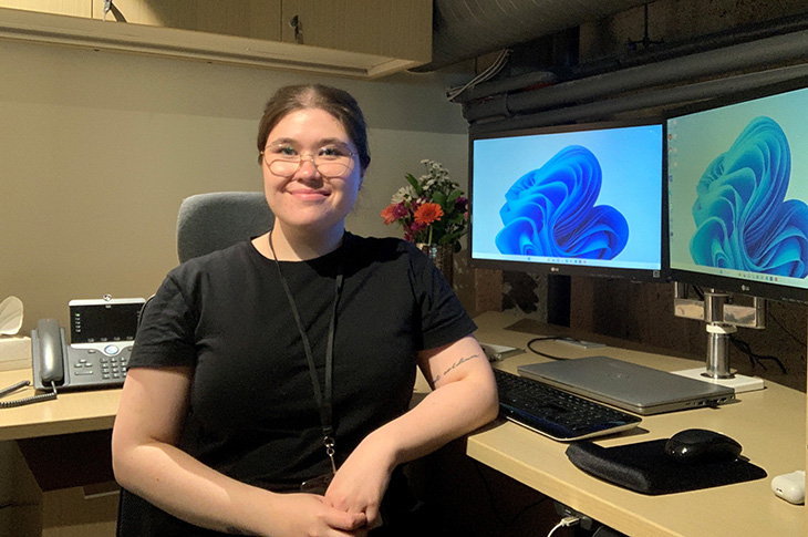 Veronika sits at her desk smiling with two computer screens in the background. 