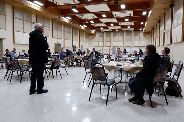 Person speaks while holding a microphone in front of a room full of people sitting around tables.