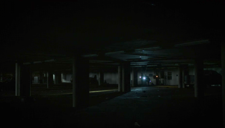A distance shot of Joel, Ellie, Sam and Henry holding flashlights and looking around in a very dark parkade. Cement pillars can just be made out in the dark space.