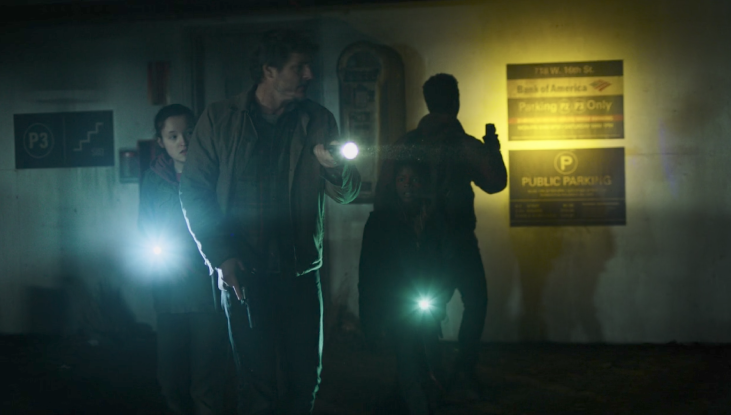 A close shot of Joel, Ellie, Sam and Henry holding flashlights and looking around in a very dark parkade.