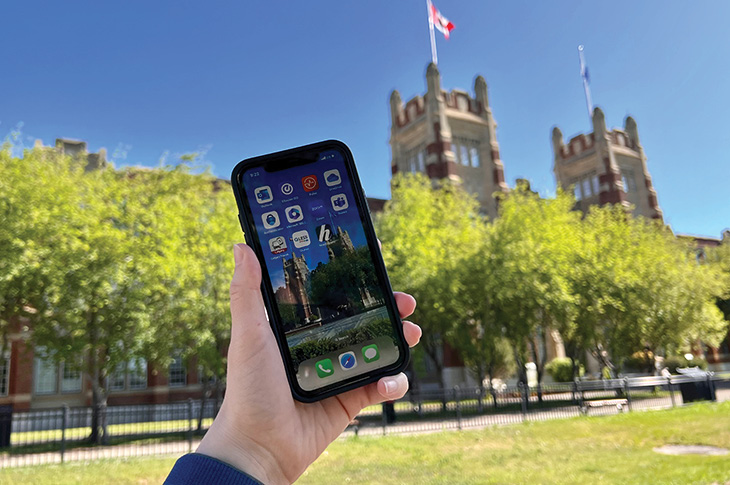 A phone being held in front of Heritage Hall on a sunny day with the recommended apps on the screen.