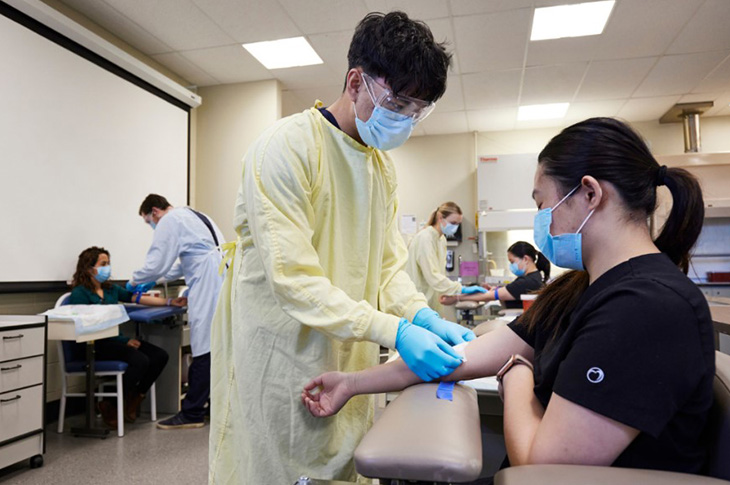 A student in a medical gown, gloves and mask tapes another student’s arm in a medical lab classroom.