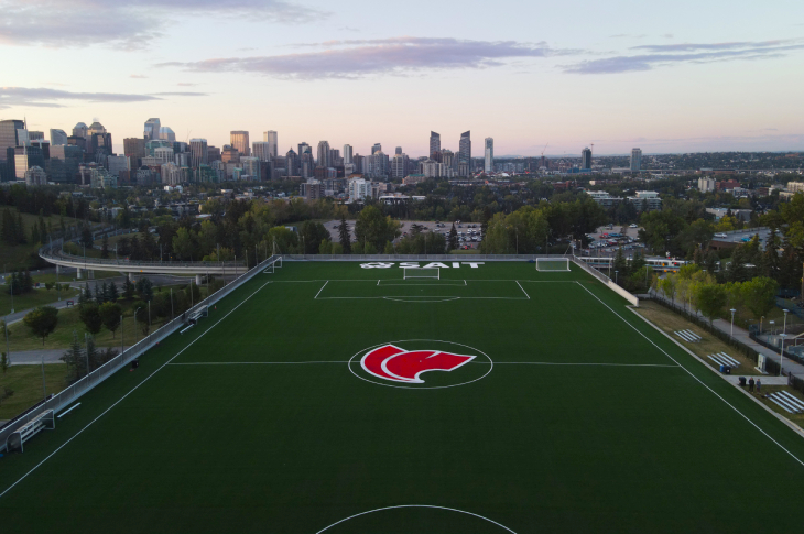 An aerial photo of SAIT's Cohos Field, a soccer-field with green astroturf and the Trojans logo in the middle. Behind the field is a view of the Calgary skyline at sunrise.
