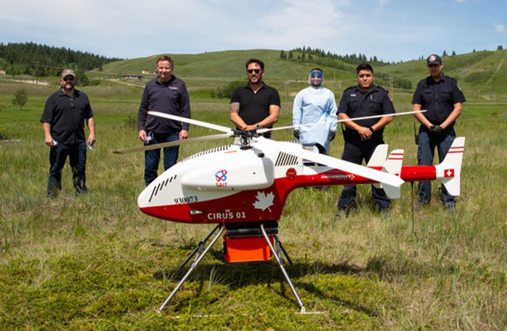 A group of six people stand in a field behind a drone that is red and white, approximately four feet tall and is similar to a small helicopter. 