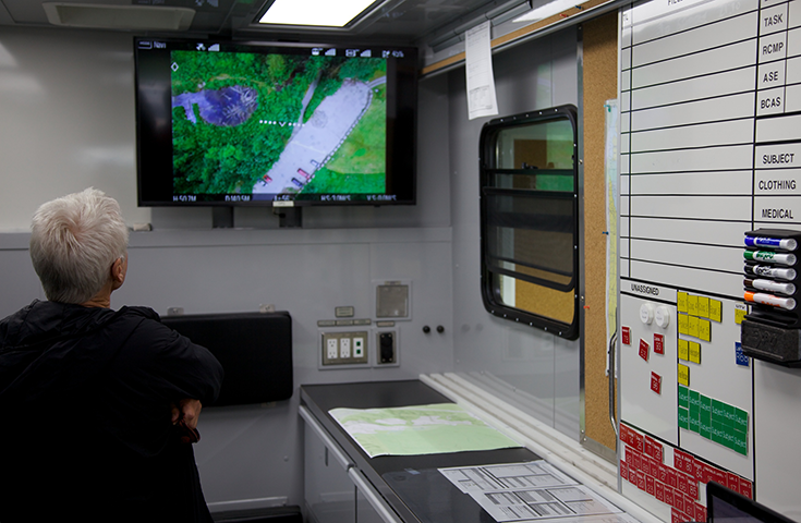 A man stands in a trailer looking at a TV screen that shows a forest from above.