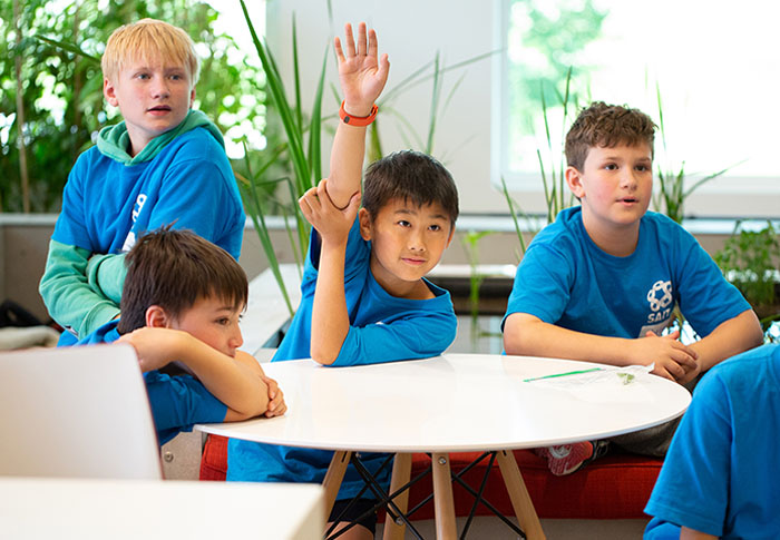 Four young SAIT campers sit around a table. Their focus is past the camera. One of the students is raising their hand to ask a question.