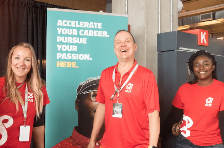 Three SAIT employees stand at a booth at SAIT's Open House. They are in front of a sign that says "Accelerate your future. Pursue your passion. Here."