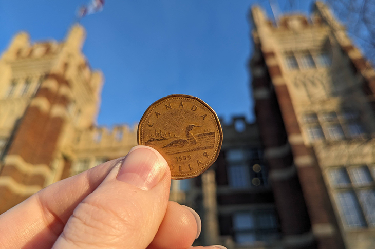 A hand holding a loonie in front of SAIT's Heritage Hall